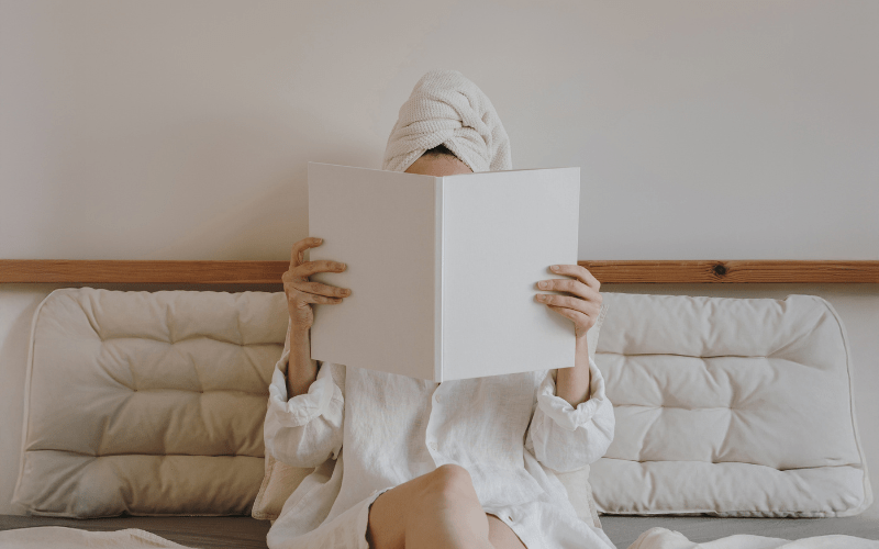 A woman relaxing on a bed with a hair towel wrap reading a book, representing a peaceful in-room spa afternoon by Lumi Glow Bangkok.