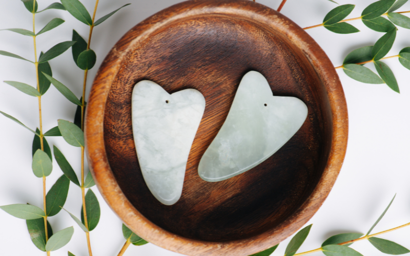 Two jade Gua Sha scraper tools in a wooden bowl with eucalyptus leaves, used for facial massage delivery treatments by Lumi Glow Bangkok.